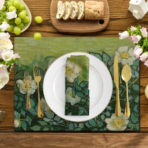 Può includere: Un runner da tavolo verde e bianco con un design floreale con rose bianche e foglie verdi. Il runner è apparecchiato con un piatto bianco, posate d'oro e un tovagliolo bianco piegato con un design floreale.