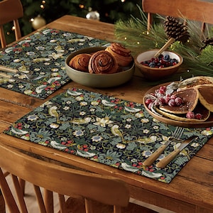 Peut inclure: Deux sets de table avec un fond vert foncé et un motif floral avec des oiseaux et des baies. Les sets de table sont sur une table en bois avec un bol de pâtisseries, un bol de baies et une assiette de crêpes.