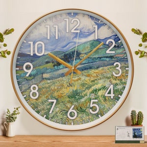 May include: A round wall clock with a gold frame and white face. The clock face features a painting of a landscape with a blue sky and green hills. The numbers are white and the hands are gold.
