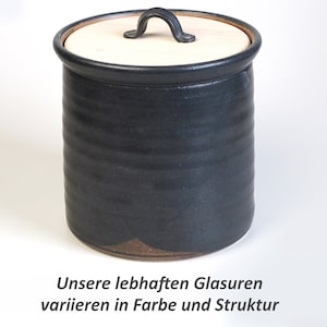 Könnte beinhalten: Eine schwarze Keramikdose mit einem Holzdeckel und einem schwarzen Metallgriff. Die Dose hat unten einen braunen Überzug. "Unsere lebhaften Glasuren variieren in Farbe und Struktur" steht in schwarzer Schrift unten im Bild.