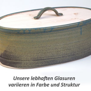 Puede incluir: Caja de pan ovalada de cerámica con tapa de madera y asa de metal. La caja tiene un esmalte verde y marrón con un acabado texturizado. "Unsere lebhaften Glasuren variieren in Farbe und Struktur" está escrito en alemán debajo de la caja.