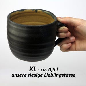 Könnte beinhalten: Eine große, schwarze Keramiktasse mit braunem Innenraum und einem abgerundeten Henkel. Die Tasse ist mit "XL - ca. 0,5 l unsere riesige Lieblingstasse" gekennzeichnet.