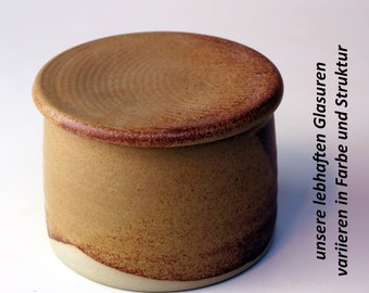 Può includere: Un porta burro in ceramica con coperchio. Il porta burro è di colore marrone chiaro con una glassa marrone più scura. Il coperchio ha un bordo leggermente rialzato.