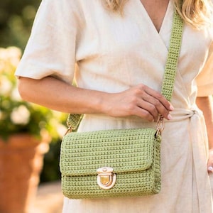 Puede incluir: Un bolso bandolera de ganchillo verde claro con forma rectangular y cierre dorado. El bolso tiene una correa de ganchillo a juego y se lleva al hombro. El bolso mide aproximadamente 23 cm de ancho y 15 cm de alto.