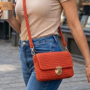 May include: An orange crochet crossbody bag with a gold-colored clasp and braided strap. The bag is rectangular with a flap closure. The person is wearing blue jeans and a tan t-shirt. A stainless steel travel mug with "CITY CAFE" is held in the hand.