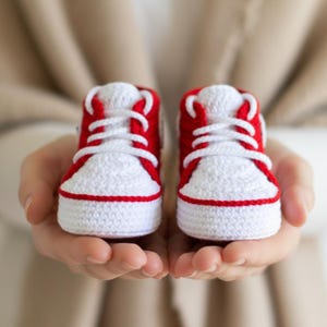 May include: A pair of handmade, crocheted baby sneakers. The shoes are white with red accents on the toe, heel, and trim. White laces are tied in a crisscross pattern. The sneakers are held in open hands.