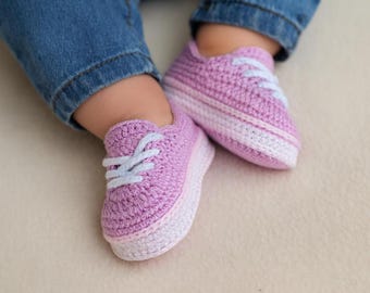 May include: A pair of handmade, crochet baby sneakers in a soft lavender color. The shoes feature white laces and a white stripe along the sole. The baby is wearing blue jeans.