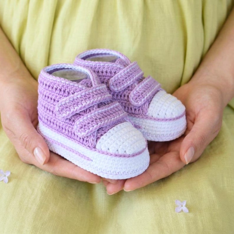 May include: A pair of handmade crochet baby sneakers in lavender and white. The high-top shoes feature hook-and-loop closures and a white toe cap. The shoes are held in open hands against a yellow-green background.