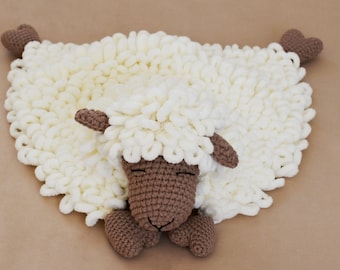 May include: A handmade, cream-colored sheep-shaped blanket. The blanket features a fluffy, looped texture, with a crocheted brown head, ears, and feet. The sheep's face has closed eyes, suggesting a sleeping or relaxed pose. The blanket is designed for babies.