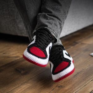 May include: A pair of handmade crochet trainers featuring a colour-blocked design in red, white, and black. The shoes have a rounded toe box, black laces, and a thick white sole, mimicking the appearance of high-top athletic footwear in yarn.