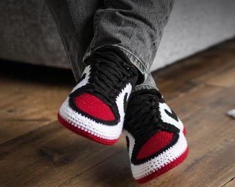 May include: A pair of handmade crochet sneakers featuring a color-blocked design in red, white, and black. The shoes have a rounded toe box, black laces, and a thick white sole, mimicking the appearance of high-top athletic footwear in yarn.