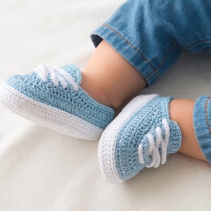 Può includere: Un paio di sneakers all'uncinetto azzurre e bianche con lacci bianchi. Le scarpe sono realizzate con un filato morbido e strutturato e hanno un classico design da sneaker. Le scarpe sono mostrate su una superficie bianca.