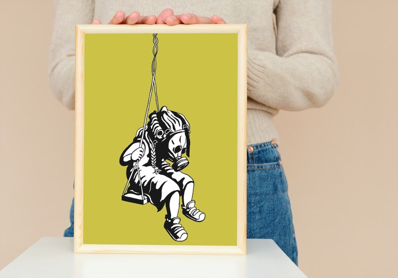 May include: Black and white artwork of a child wearing a gas mask, swinging on a swing. The artwork is framed in a light wood frame and set against a yellow-green background. The image is a print.