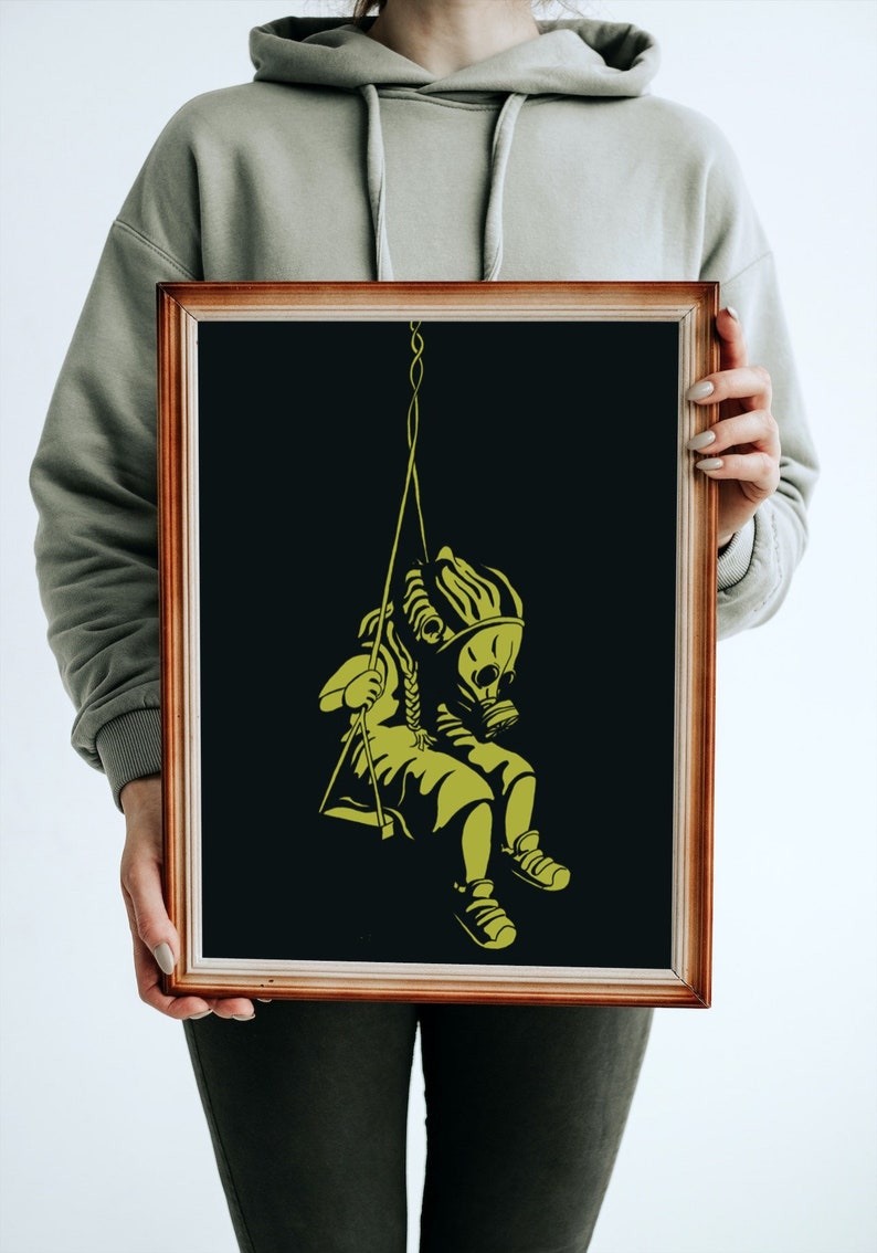 May include: Framed art print featuring a yellow illustration on a black background. The artwork depicts a figure wearing a gas mask and sitting on a swing. The frame is a warm brown color.