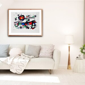 May include: A framed abstract painting with black lines, red, blue, and green shapes on a white background. The painting is hanging on a white wall above a white couch with pillows and a throw blanket.
