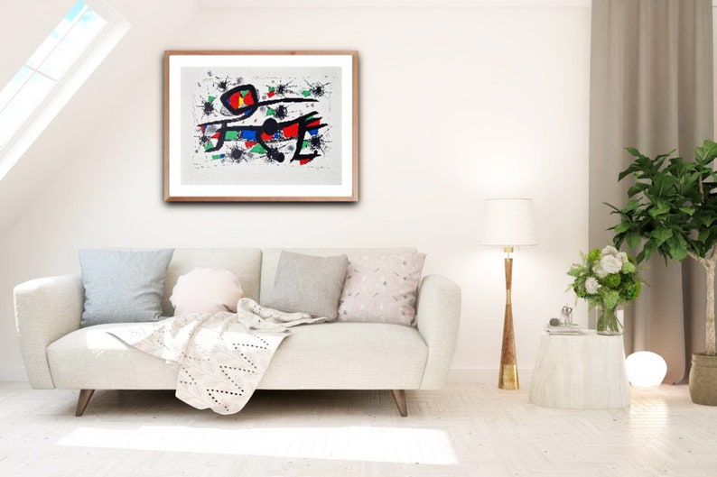 May include: A framed abstract painting with black lines, red, blue, and green shapes on a white background. The painting is hanging on a white wall above a white couch with pillows and a throw blanket.
