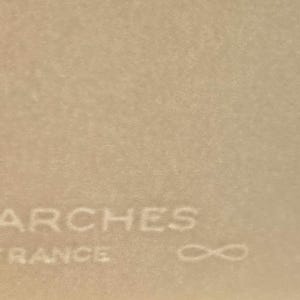 May include: Close-up of a textured, beige paper surface. The word "ARCHES" is embossed in the upper portion, with "FRANCE" below it. An infinity symbol is to the right of "FRANCE".