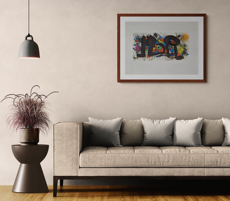 May include: Abstract art print in a dark wood frame. The artwork features a colorful, abstract design with black, blue, orange, and yellow shapes. The print is mounted on a white background and hangs above a beige sofa.