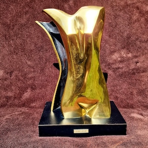 Könnte beinhalten: Eine polierte, goldfarbene abstrakte Skulptur mit glatter, geschwungener Form und einem dunklen Innenraum. Die Skulptur ist auf einem rechteckigen schwarzen Sockel montiert, der ein kleines weißes Etikett mit dem Text C.10 150 in schwarzer Schrift trägt.