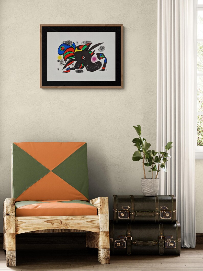 May include: Abstract art print in a wooden frame. The artwork features a black figure with colorful accents of red, blue, green, and yellow. A wooden chair with orange and green cushions is in the foreground, with a stack of dark brown vintage suitcases.