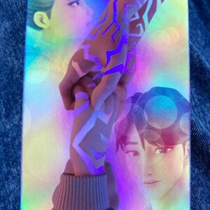 May include: Holographic trading card featuring two anime-style characters holding hands, with a glowing purple design on the arm. The card has a rainbow iridescent finish, with soft focus portraits in the background.