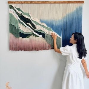 May include: A large wall hanging made of fiber, suspended from a wooden rod. The piece features a gradient of colors including cream, sage green, dark forest green, and a deep blue ombre effect, with a soft pink hue at the bottom edge.