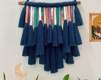 Pode incluir: Uma tapeçaria de macramé azul com uma barra de madeira na parte superior. O macramé tem uma franja em camadas com borlas em tons de rosa, branco, turquesa e verde.
