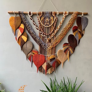 May include: A large macrame wall hanging with a wooden dowel at the top. The macrame is made with a variety of natural coloured cords and features a variety of knots and patterns. The macrame is decorated with wooden beads and has a fringe at the bottom. The macrame is hanging on a light grey wall. "LUNAR MACRAME STORE" is written in orange text at the bottom of the image.