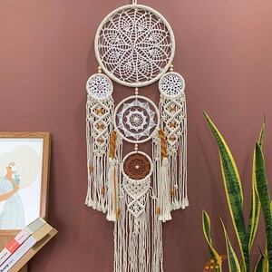 May include: A white and brown macrame dream catcher with multiple layers of intricate knotting and beads. The dream catcher is hanging on a brown wall with a green plant in the background.