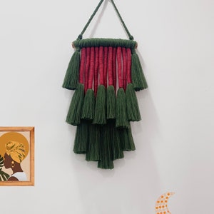 May include: A green and red macrame wall hanging with tassels. The hanging is made of cotton cord and is attached to a wooden dowel. The hanging is in the shape of a triangle and has a fringe at the bottom. The hanging is a popular home decor item.