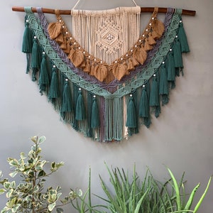 Puede incluir: Un gran tapiz de macramé para colgar en la pared con un diseño geométrico en tonos beige, verde y morado. El tapiz presenta una forma de diamante central con borlas y cuentas.