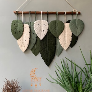 May include: Macrame leaf wall hanging with a wooden dowel and a selection of green and off-white leaves. The leaves are arranged symmetrically, creating a natural and decorative element. The wall hanging is suspended by a natural cord.