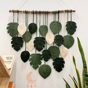 May include: A macrame wall hanging featuring a design of green and white leaves. The leaves are made of yarn and are hanging from a wooden branch. The wall hanging is on a white wall with a green plant in the corner.