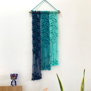 May include: A wall hanging made of macrame with a blue, teal, and turquoise color gradient. The macrame is made of cotton cord and is hung on a wooden dowel rod. The wall hanging is a modern and stylish way to add a touch of bohemian chic to any room.