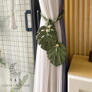 May include: Green macrame curtain tiebacks in a leaf design with wooden beads. The tiebacks are holding back white and gray curtains. A macrame hanging decoration is visible on the left. The text "LUNAR MACRAME STORE" is at the bottom.