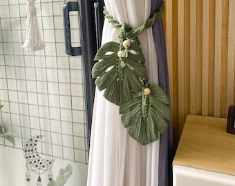 May include: Green macrame curtain tiebacks in a leaf design with wooden beads. The tiebacks are holding back white and gray curtains. A macrame hanging decoration is visible on the left. The text "LUNAR MACRAME STORE" is at the bottom.