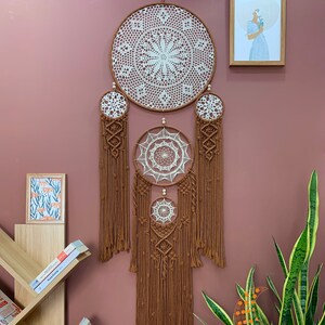 May include: A large brown macrame dream catcher with three white crocheted circles. The dream catcher is hanging on a pink wall with a small framed print and a potted plant.