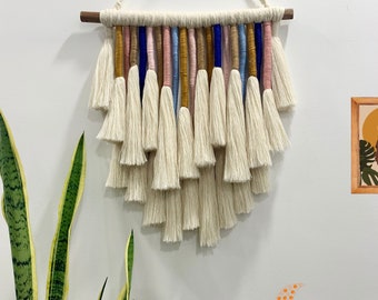 Pode incluir: Uma tapeçaria de macramé branca com uma barra de madeira na parte superior. O macramé apresenta uma cascata de franjas com fios coloridos tecidos, incluindo azul, rosa e amarelo.