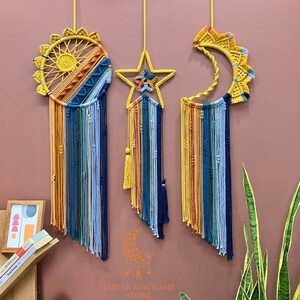 Op de afbeelding: Drie macramé wandhangers in de vorm van een zon, een ster en een halve maan. Elke hanger heeft een regenboog aan kleuren, waaronder geel, oranje, rood, blauw, groen en paars. De hangers zijn gemaakt van katoenen koord en hebben kwastjes aan de onderkant.