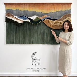 以下が含まれることがあります： 風景デザインの大きなテクスチャードウォールハンギング。このアートワークは、オレンジ、グレー、グリーン、ブラウンなどの色のグラデーションを特徴とし、上部に木の棒があります。下部には長い緑色のフリンジがあります。このアートワークは白い壁に展示されています。