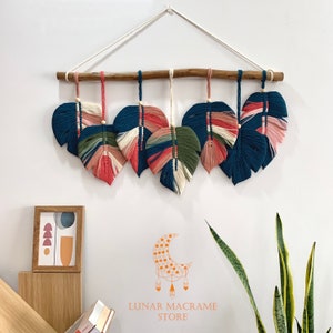 May include: A macrame wall hanging with six leaf-shaped pieces in shades of blue, pink, green, and white. The leaves are attached to a wooden dowel and hang from a white cord.