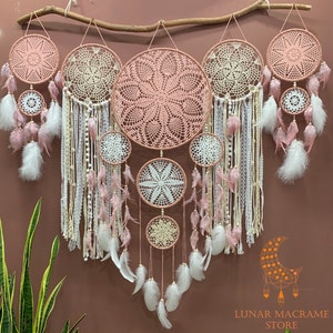 May include: A set of six pink and white dreamcatchers with feathers hanging from a wooden branch. The dreamcatchers are made of crocheted lace and have a variety of sizes. The largest dreamcatcher has a pink crocheted center and is surrounded by a white crocheted border. The other dreamcatchers are smaller and have a variety of designs. The dreamcatchers are all hanging from a wooden branch and have feathers hanging from them.