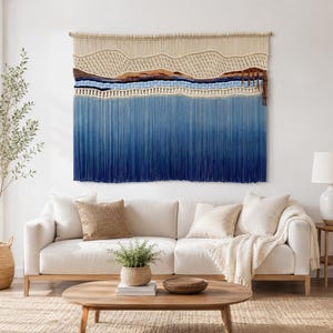 May include: A large macrame wall hanging featuring a landscape design with cream-colored woven patterns at the top, followed by horizontal bands of brown, black, and light blue, transitioning into a long, dense fringe of ombre blue cotton cords.