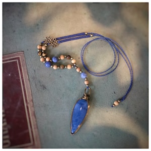Puede incluir: Un collar con un colgante de piedra azul, una hebra de cuentas de madera y cuentas de ojo de tigre. El colgante está envuelto en alambre y cuelga de un cordón azul.