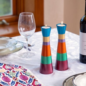 May include: Two colorful candlesticks with a red, orange, green, and blue striped design. The candlesticks are on a white tablecloth with a wine bottle and a glass of water in the background.