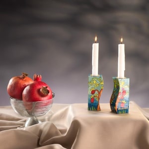 Puede incluir: Dos velas blancas en candelabros pintados a mano en azul y verde con diseños de granada. Un cuenco de granadas rojas se encuentra sobre un mantel beige.