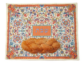 May include: A braided loaf of bread with sesame seeds rests on a decorative embroidered mat. The mat features a floral design in shades of orange, blue, and green, with Hebrew text in a rectangular frame.