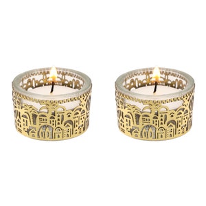 May include: Two lit votive candles in clear glass holders with gold-colored, decorative metal sleeves. The sleeves feature a cut-out design of buildings and arches. The candles are white, and the flames are bright yellow.