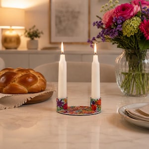 May include: A floral-patterned candle holder with two lit white candles. A round challah bread sits on a wooden plate to the left, and a vase of flowers is on the right. The table is white.