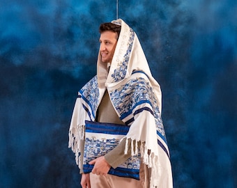 Puede incluir: Un hombre lleva una chal de oración blanca con bordados azules y dorados. La chal tiene flecos y está decorada con un patrón de edificios. Está sosteniendo un libro de oraciones azul y blanco.